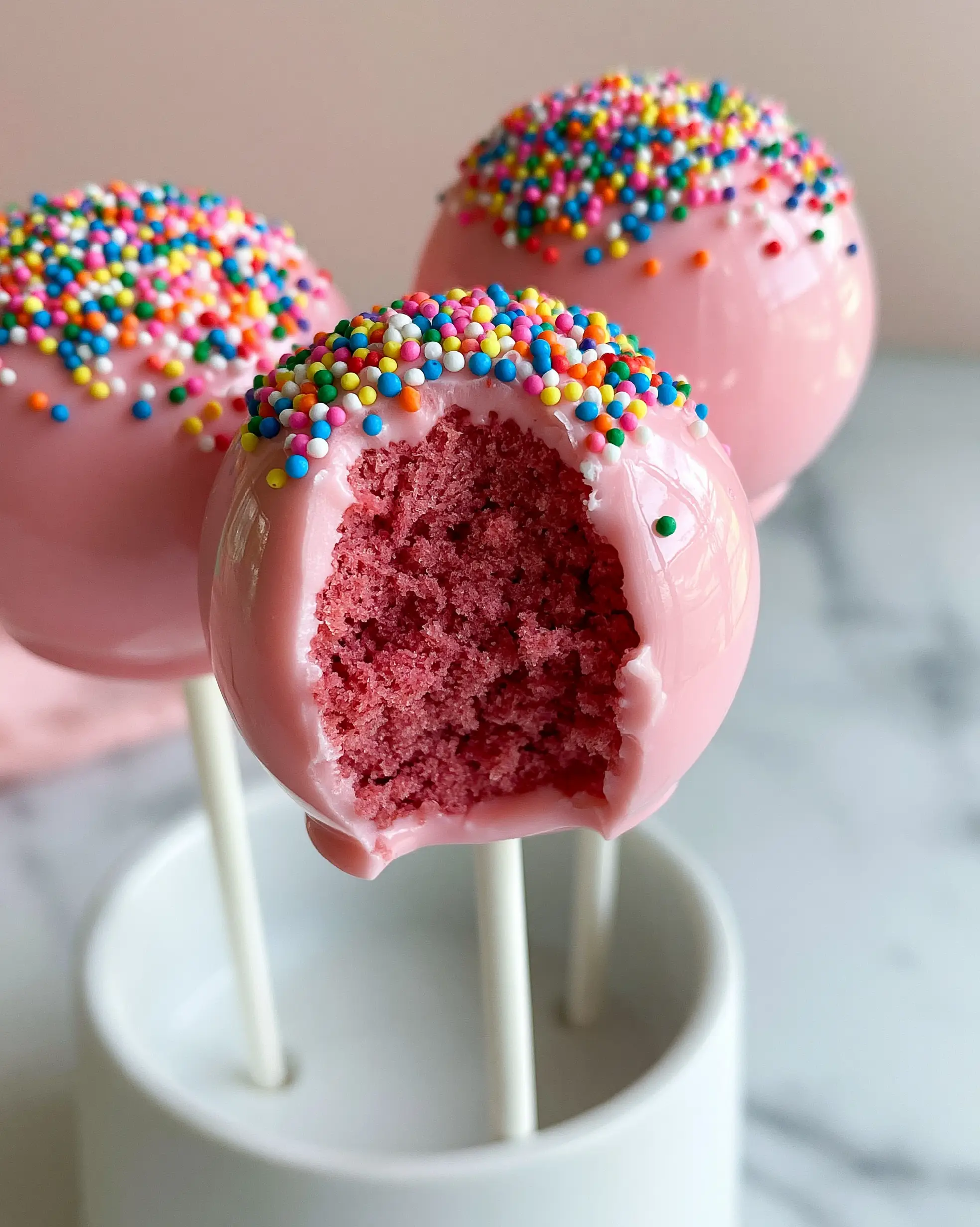 Handheld photo of three Birthday Cake Pops, one with a bite revealing a moist pink center.